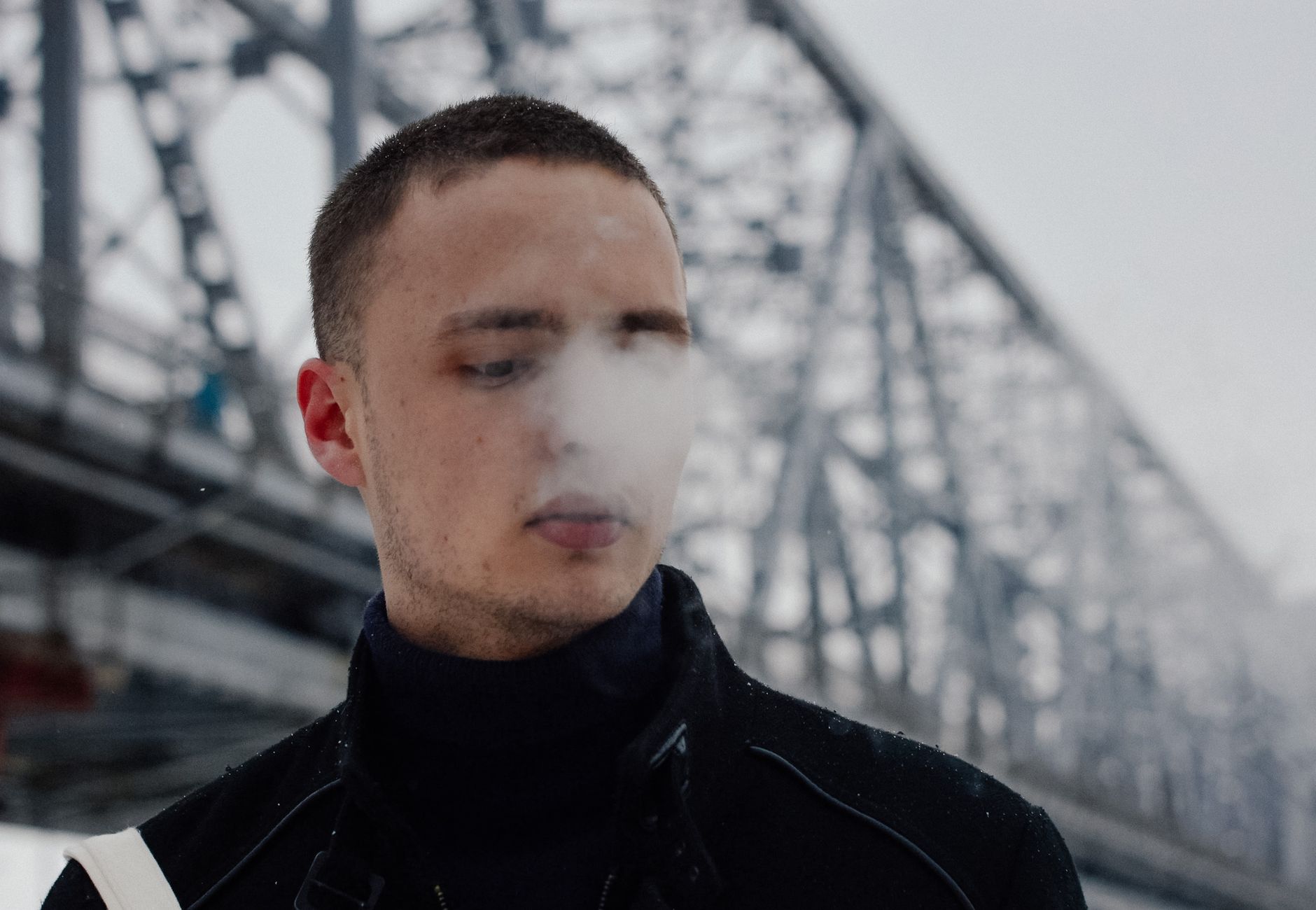 A young man exhales steam on a cold winter day, standing near a large iron bridge.