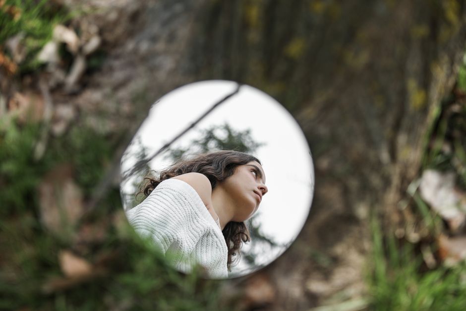 Woman's reflection in a mirror outdoors, surrounded by grass and tree trunk.